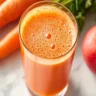 A glass of freshly poured carrot and apple juice with fresh ingredients beside it on a white marble kitchen counter.